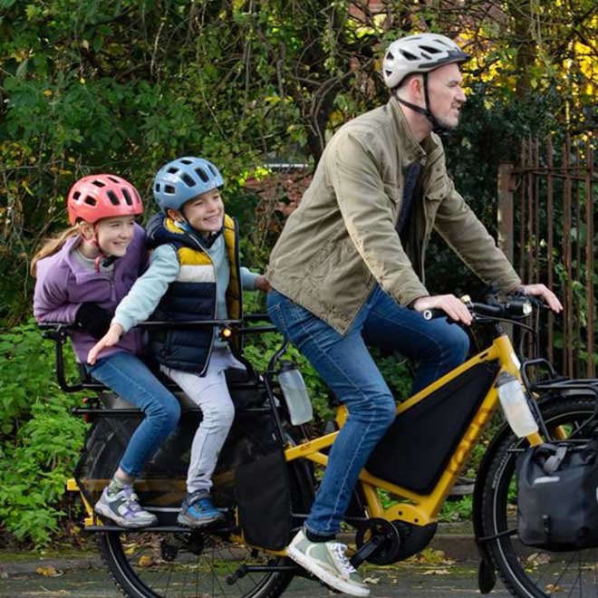 A dad riding a Tern Orox cargo e-bike with two young kids on the child seats with full passenger-carrying accessories setup. 