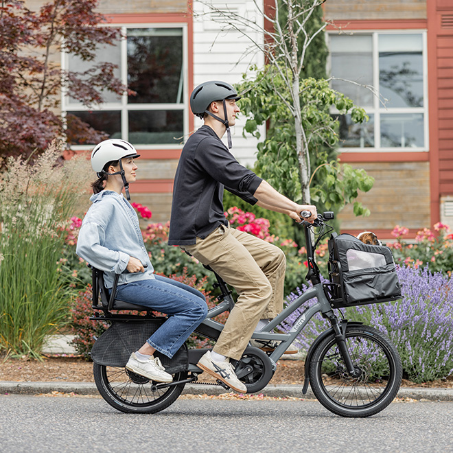 A woman riding a Tern HSD cargo e-bike with a man on the passenger seat and a bag on the front rack. 