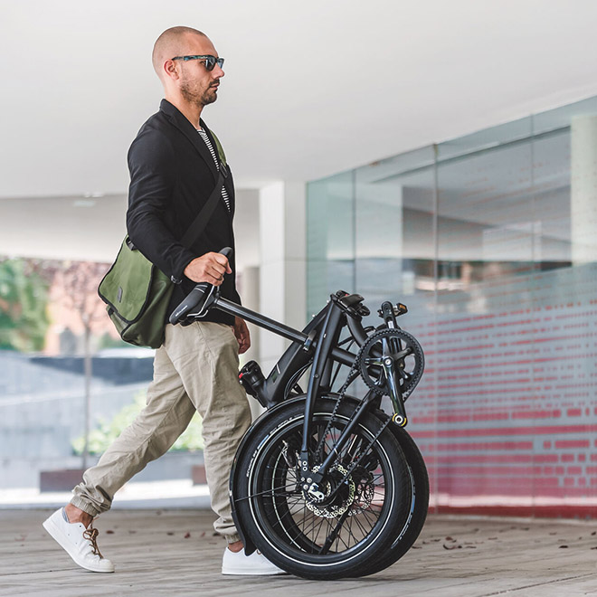 A man walks through a indoor space while carrying a folding bike by its seat post.
