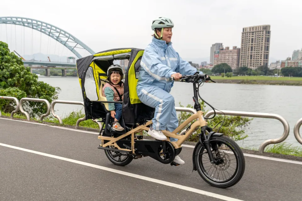 A mom riding a Tern GSD cargo e-bike on a riverside bike path in the rain, carrying a child under a rear rain canopy.